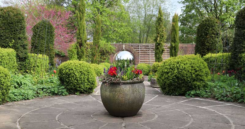 Flower pot and silver orb sit in the middle of the image with trees, shrubs and the garden wall in the background