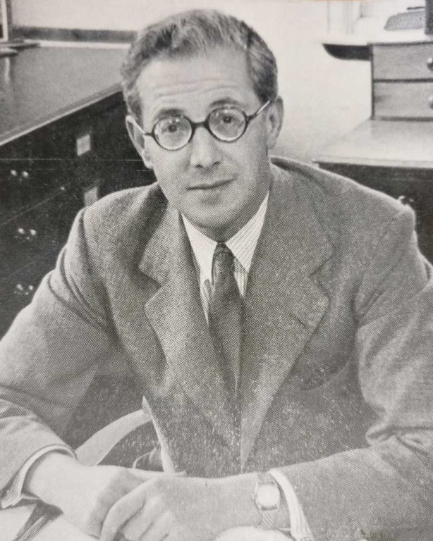 Lord Kenyon sits at a desk looking directly at the camera. The photo is black and white. Lord Kenyon is wearing a suit and tie, and has round spectacles.