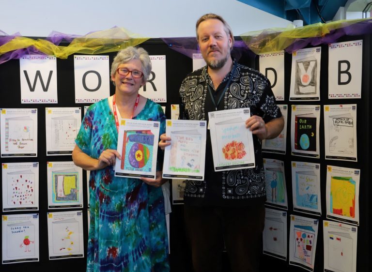 Local author Jan Hedger and functional skills teacher Iain Evans hold up the winning Book Cover Competition entries, stood in front of a World Book Day display of all the colourful entries.