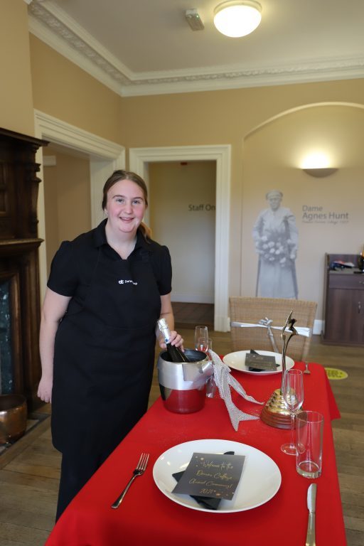 Supported intern Maisy  stands next to a restaurant table that she has laid out for a table-laying competition.