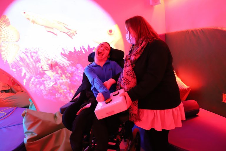 Student Jake, who has PMLD, sat in his wheelchair accompanied by PMLD lead Bev in the sensory room, which is lit up in red.