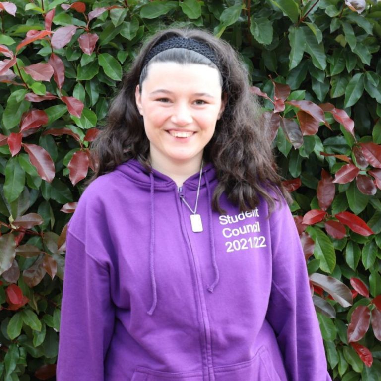 Amy pictured smiling, wearing her purple Student Council hoodie, and standing in front of a green and red shrub.