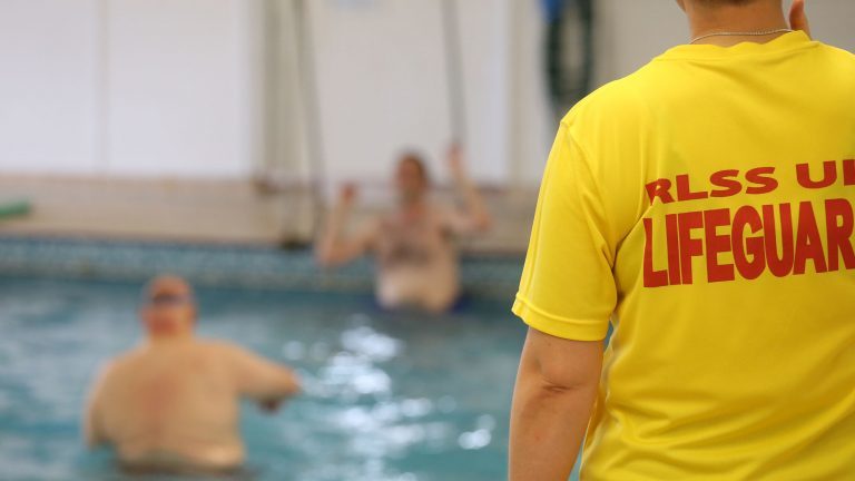 Swimming pool lifeguard has her back to the camera. Two people in the pool in the background.