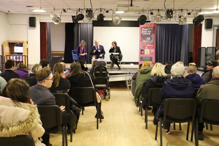 Hazel, Richard and Derwen press officer Eluned sit on the stage with a large banner featuring Richard's book next to them. A large audience are sat in chairs facing them.