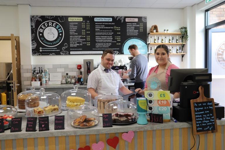 George working behind the counter - brimming with cakes - at Refresh cafe with staff member Ellie (with Grant in the background making a coffee).
