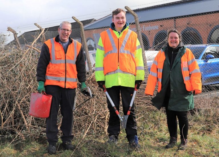 Cambrian Heritage Railways Volunteer Coordinator Phil carrying a bucket, Derwen student Harry carrying shears and teacher Ruth, all wearing orange high-vis jackets and ready for work.