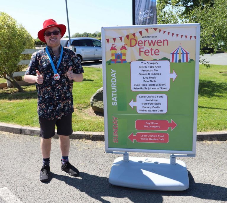 Former student, wearing a medal, and dressed in sun hat, sunglasses and tropical shirt, stands next to the large Derwen Fete banner which lists all the events for the weekend.