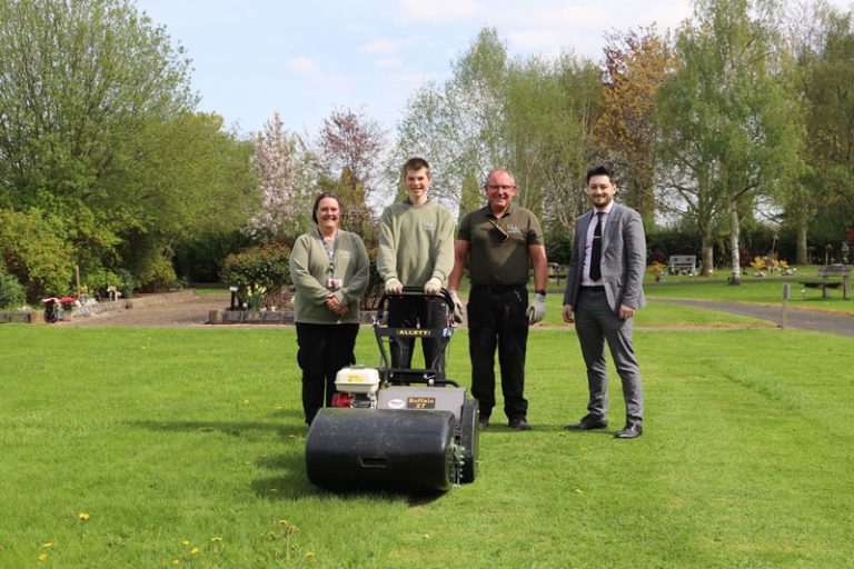 Teacher Ruth, student Oliver, grounds maintenance Peter (dressed in green sweatshirts) and business lead Kristen (dressed in suit) stand together on the lawn behind the industrial lawnmower.