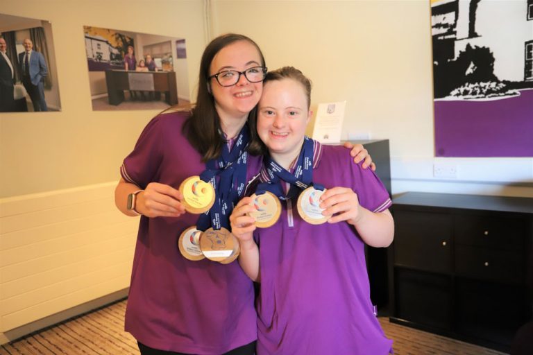 Students Helen and Lizzie wearing purple Premier Inn tops and with medals hug each other in the Hotel 751 voyer.