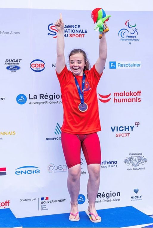 Helen, wearing red t-shirt and shorts and flipflps, holding the Virtus Global Games masoct and wearing her Gold medal, cheers with her arms in the air.