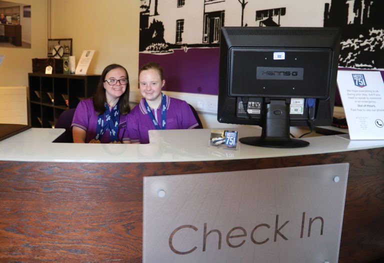 Derwen College students Helen and Lizzie - wearing purple Premier Inn polo shirts - sit behind the Check In desk in the College's Premier Inn styled Hotel 751.