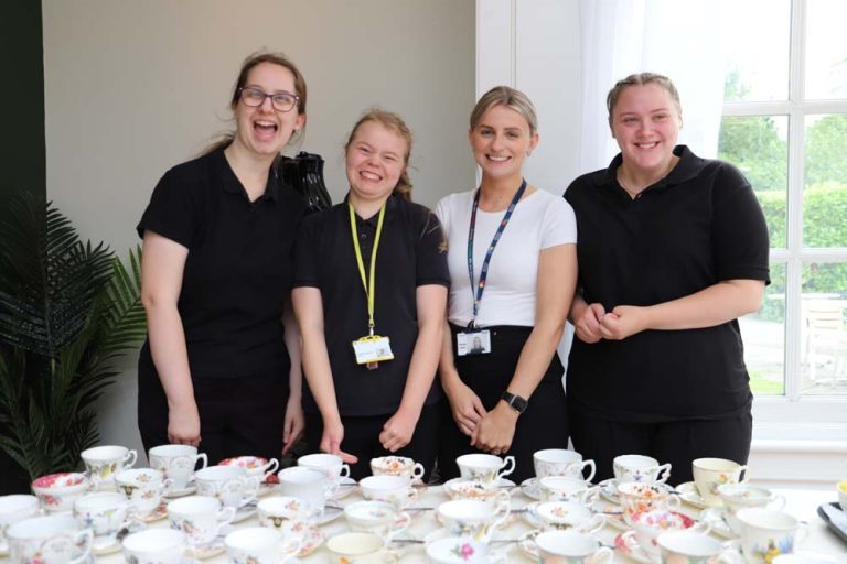Three Hospitality and Food students, dressed in black uniforms, with their Hospitality teacher, stood smiling behind a table of floral cups and saucers.