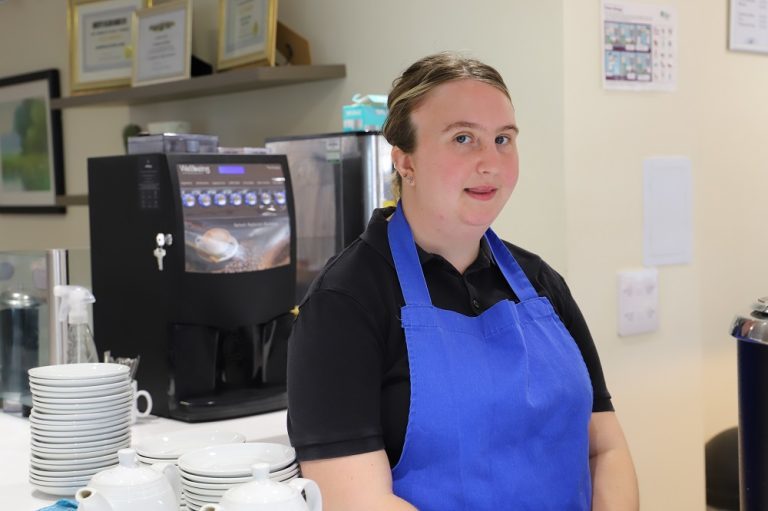 Head and shoulders image of Maisy with hair tied back and wearing blue apron, stood behind the cafe counter in front of the coffee machine.