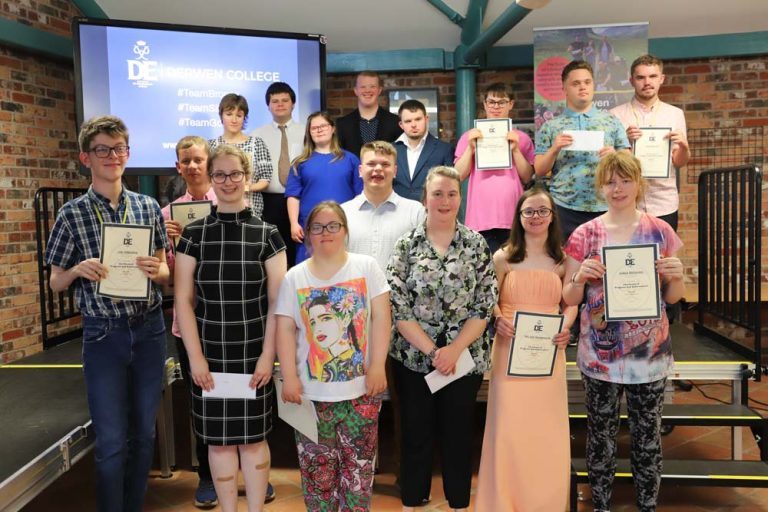 Group shot of 16 students holding Gold DofE certificates, stood on a stage with the DofE logo on a screen behind them.