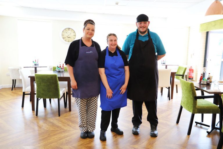 Maisy is standing among tables and chairs in the cafe, with cafe staff members stood on either side of her. They are all wearing smart aprons and smiling.