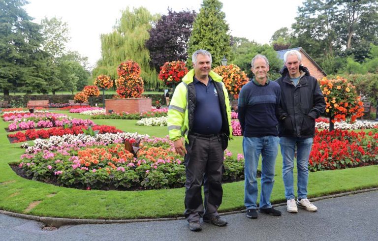 Client Anton in gardening wear and high vis jacket, nursery manager Paul Moss and client Thomas stood in front of colourful bedding plant displays in the Oswestry's park.