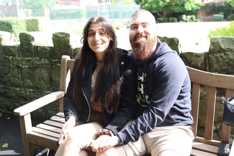 A happy couple sat on a park bench together, showing off the new engagement ring.