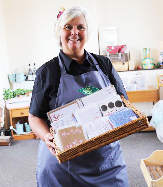 Teresa, who runs the Station Cafe, carrying a basket of greetings card to stock the pop-up shop. Teresa is wearing a branding apron with The Station Cafe logo.