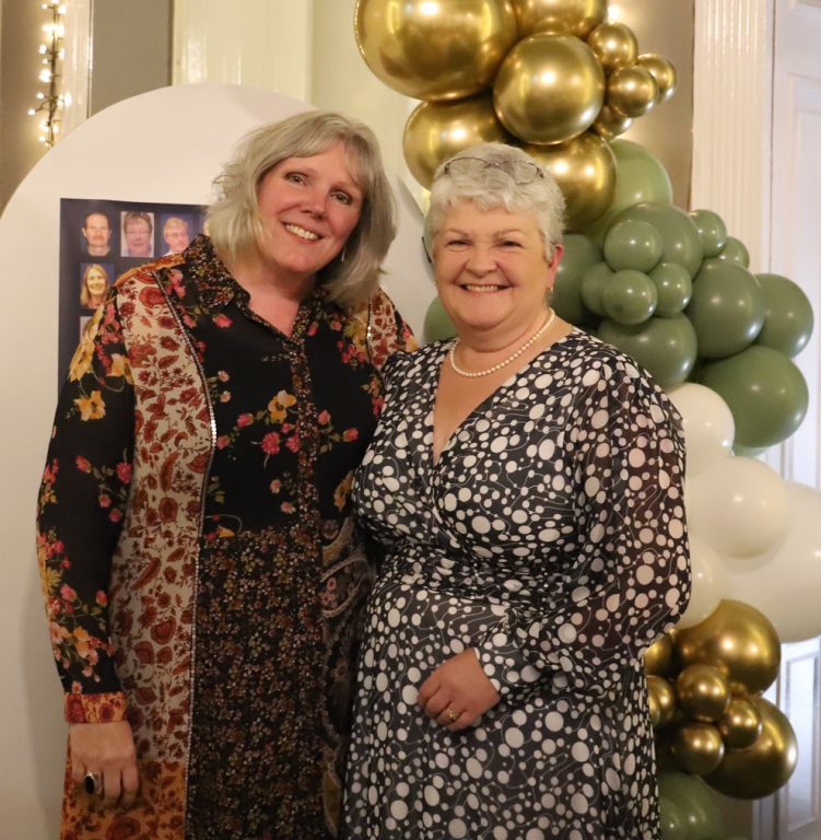 Principal Meryl stands next to Alison Thomas, both wearing smart dresses and smiling. Behind is a poster of all the long-serving staff and gold and green balloons.