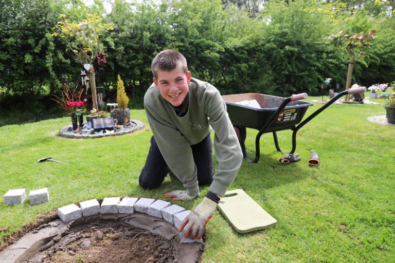 Oliver, smiling and dressed in green work sweatshirt, kneels down to lay bricks as part of his work at Shrewsbury Crematorium.