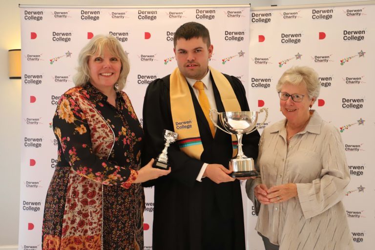 College Principal Meryl Green holding a small silver cup trophy, Student of the Year Mark (in black gown and gold Derwen College sash) and Chair of Governors K Kimber with a large silver cup trophy.