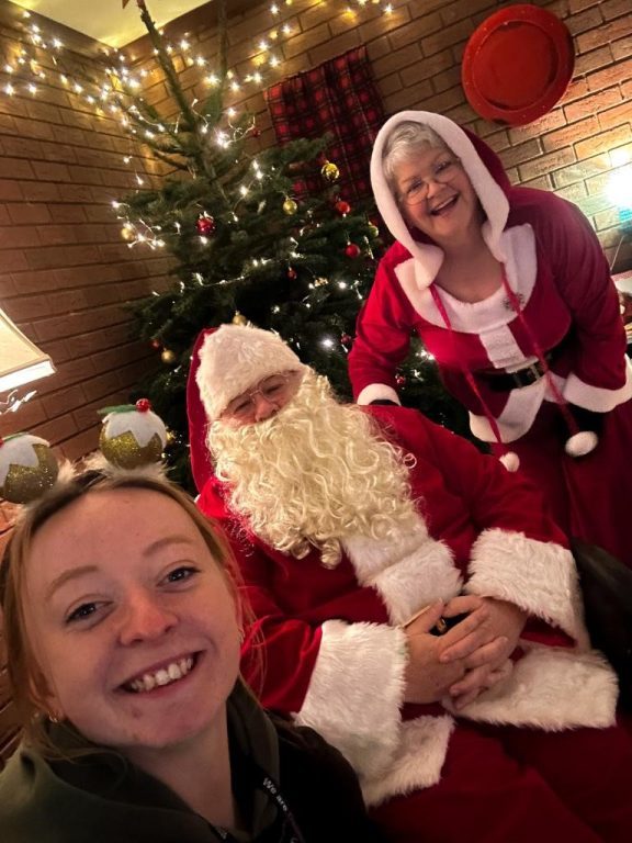Selfie of fundraiser Meg with Christmas pudding headwear, along with Santa and Mrs Christmas with Christmas tree behind.