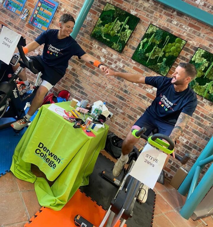 Derwen sporty spinners Kelli and Steve sat next to each other on spinbikes - wearing Derwen Charity t-shirts - share a fist pump.