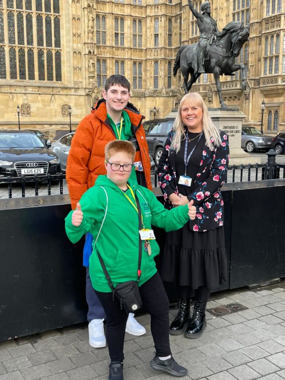 Students Byron and AJ, wearing their green Student Union Board hoodies, stand outside the Houses of Parliament with Director of Learning and Curriculum Development Zoe Wood.