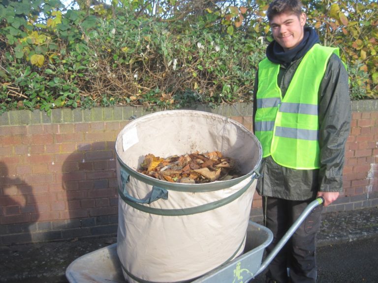 Supported intern Scott, dressed in a high-vis waistcoat, pushes a wheelbarrow containing cleared up leaves.