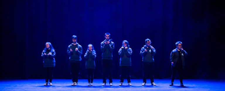 seven performers on stage standing under blue spotlights