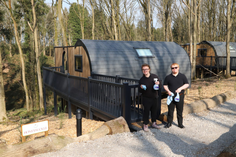 Two students dressed in black and holding cleaning spray and cloth stand in front of a woodland lodge.