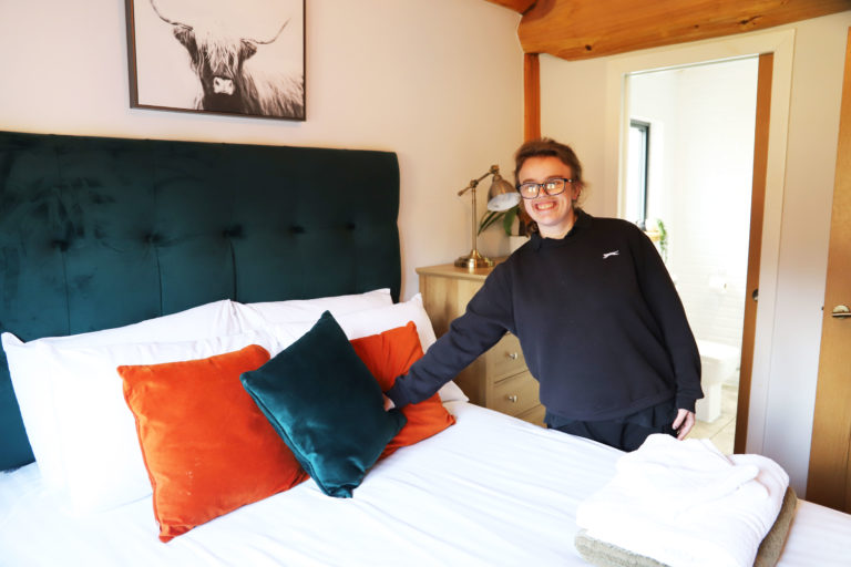 Student Brodie arranges the orange and teal cushions on the white bedding on the luxurious double bed.