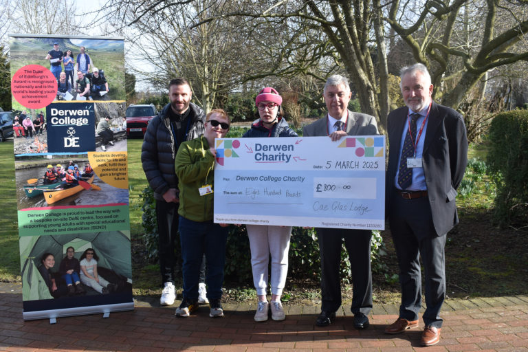 Five people stand facing the camera. Three of them hold a big cheque