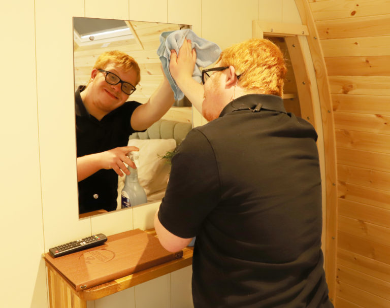 Student Tom cleans the mirror inside a wooden lodge, looking into the mirror and smiling at the camera.