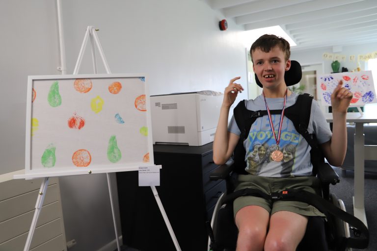 Student Ben, sat in a wheelchair, next to his artwork with fruit printed on to it.