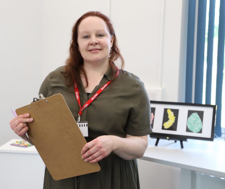 A woman holding a clipboard, stood in front of works of art, smiling.