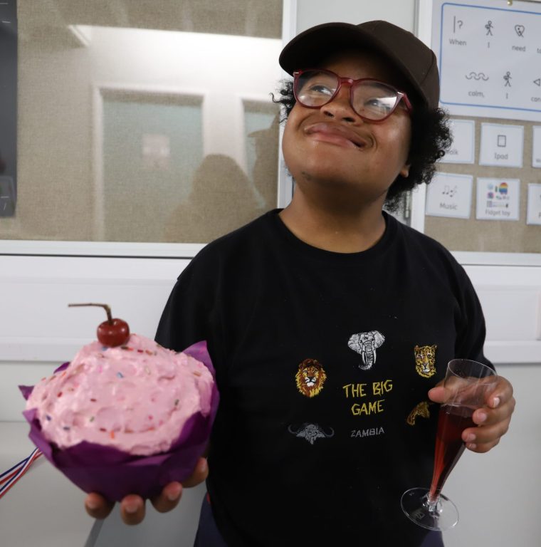 Student Joy smiling holding a huge pink cupcake made of papier mache with cherry on top, with a champagne flute of pink drink in the other.