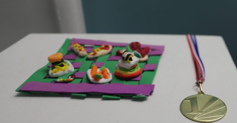 A colour picnic blanket, food and bee made out of clay with a 1st medal next to it.