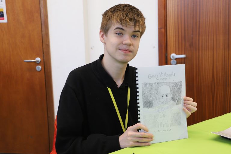 Ben sits at a desk holding up the self-illustrated front cover of Gods Vs Angels