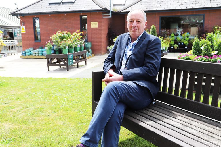 Jamie sits on a bench with plants behind him in the College Garden Centre.