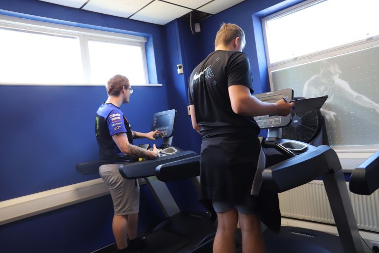 Two male students, wearing shorts and t-shirts, check the running machines in the gym.