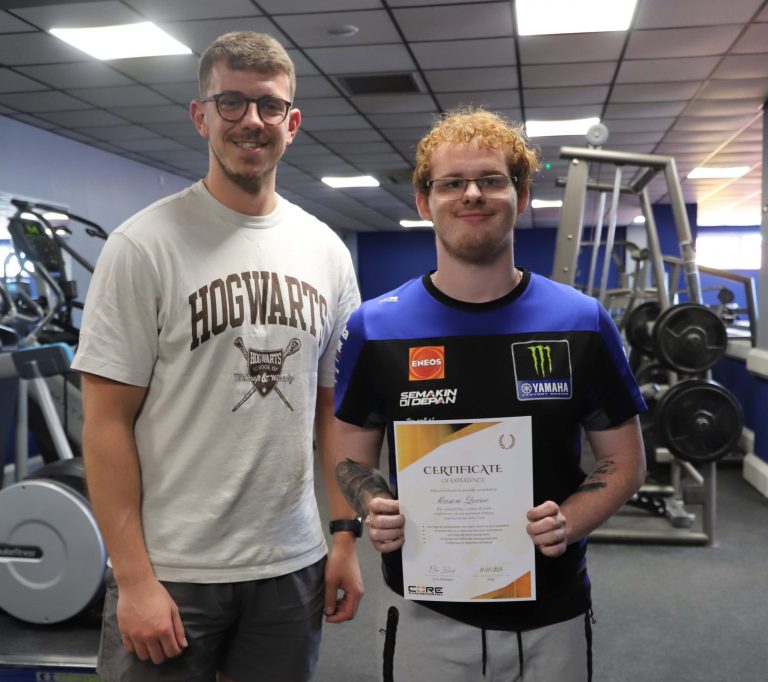 Gym owner Ollie stands next to student Mason in the gym. Mason is holding a certificate, and both are wearing t-shirts and shorts.