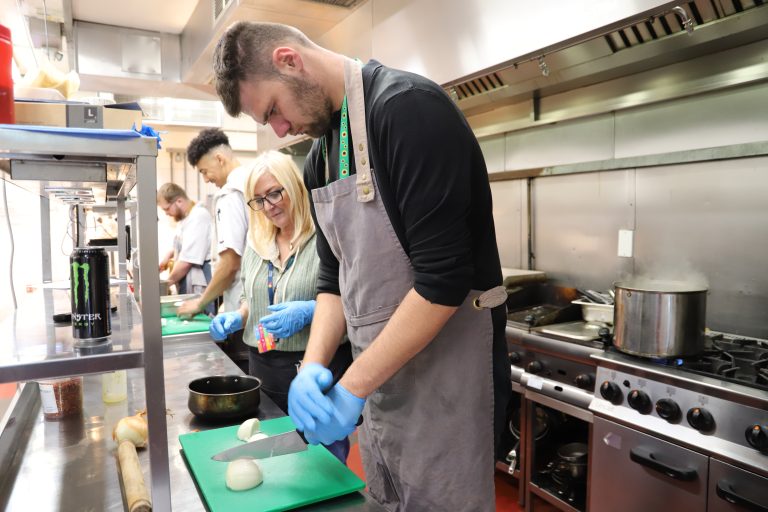 Student Josh chops an onion on a chopping board in the busy kitchen.