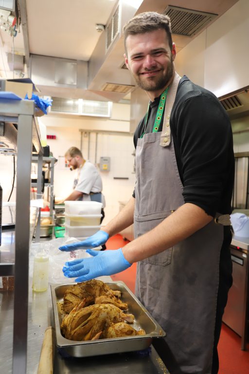 Josh, wearing an apron and blue gloves, rubs spices onto two chickens in a tray.