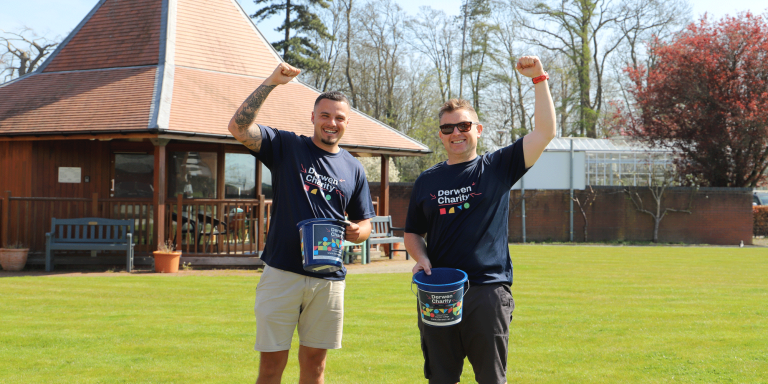 two men standing with one hand raised and one hand holding a blue charity bucket
