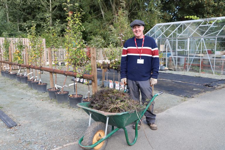Smiling man in cap and jumper with a wheelbarrow filled with weeds and prunings