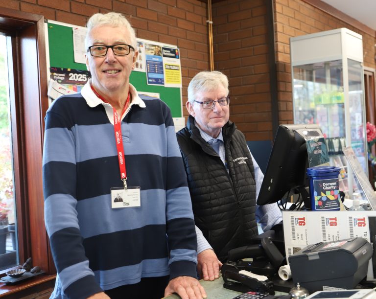 A smiling volunteer, wearing a visitor badge, behind the counter next to the till with staff member Alfie.