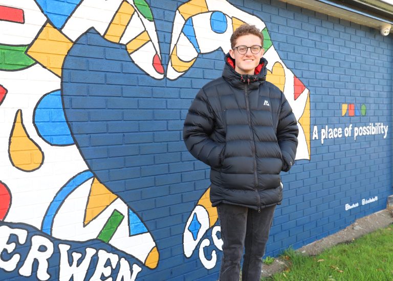 George stood outside in front of a blue painted wall with brightly coloured heart-shaped hands painted on it, as well as the name Derwen College and 'A place of possibiliity'