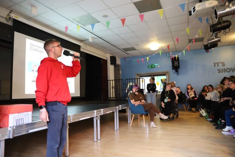 A student in a red Student Council hoodie holds a microphone to address a packed Student Union.