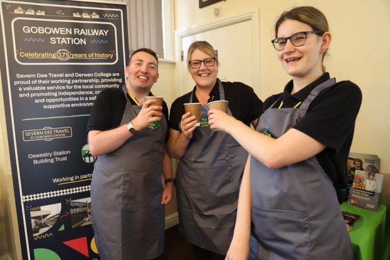 Two students and teacher, wearing Station Cafe branded aprons toast each other with cardboard coffee cups at Gobowen Railway Station